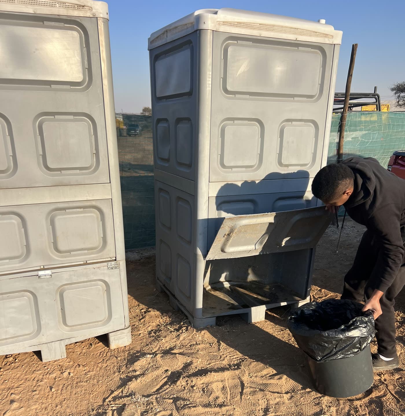 Mahapa Event Hire staff member removing the sealed waste container from the rear service hatch of a chemical toilet on a construction site in Limpopo